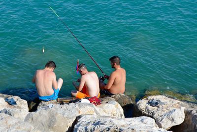 Rear view of men fishing while sitting on rock at sea