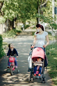 Mother wearing a mask and one daughter in a baby stroller and big sister on a bicycle pandemic 2021