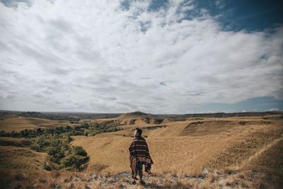 Rear view of man with blanket standing on field against cloudy sky