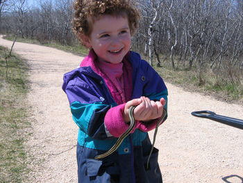 Happy boy holding garter snake while standing on road amidst bare trees