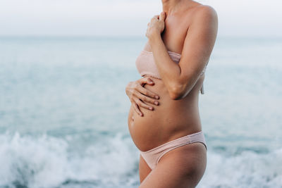 Close-up belly of a pregnant woman standing by the sea on the beach.