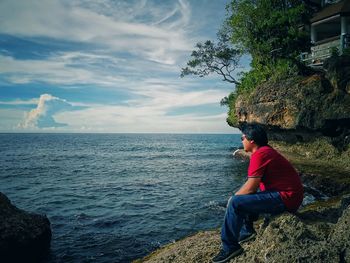 Man sitting on rock looking at sea against sky