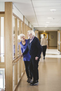 Senior couple talking while standing by window in corridor at nursing home