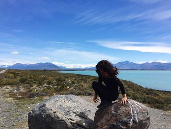 Full length of woman crouching on rock by sea against blue sky