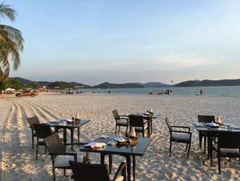 Empty chairs and tables at beach against sky