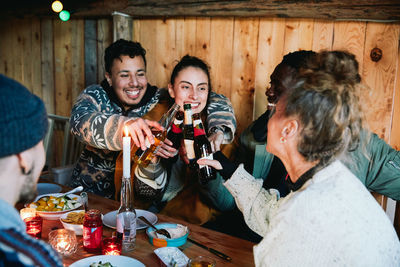 Happy male and friends toasting beer bottles while sitting in log cabin