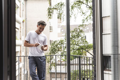 Smiling man in pyjama with cup of coffee standing on balcony