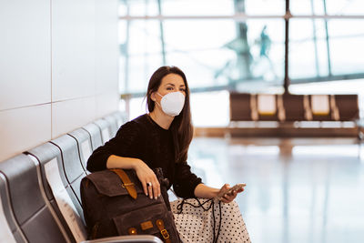 Woman wearing mask using mobile phone while sitting at airport