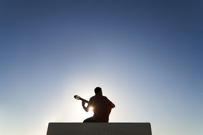 Back view silhouette of unrecognizable bearded musician playing song on acoustic guitar while sitting on border against blue cloudless sky on sunny day in city