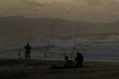 Silhouette people fishing at beach against sky