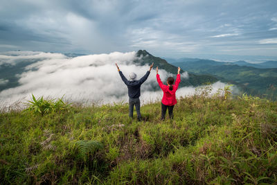 Rear view of hikers with arms raised looking at landscape against cloudy sky