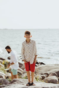 Portrait of smiling boy on rock against sea while father standing in background