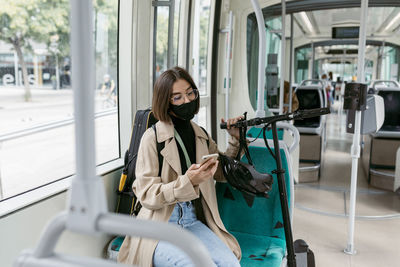 Mid adult woman wearing face mask using mobile phone while sitting with electric push scooter in tram