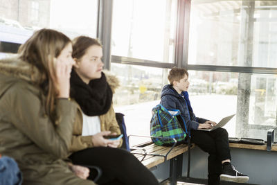 Boy using laptop with women sitting on bench in foreground at train station