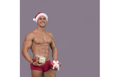 Portrait of smiling muscular man wearing santa hat while standing against gray background