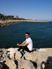 Young man sitting on rock by sea against clear sky