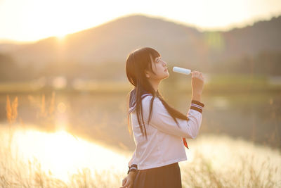 Young woman drinking water while standing against lake during sunset