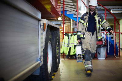 Full length of firefighter holding fire hose while walking by fire engine at station