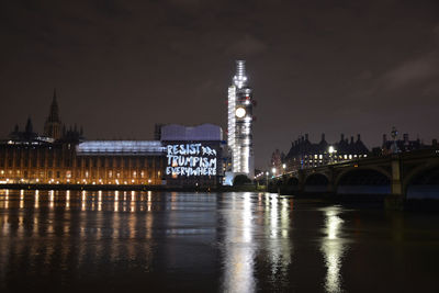 Illuminated bridge over river by buildings against sky at night