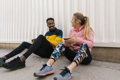Smiling male and female athletes holding mobile phone while talking with each other against wall
