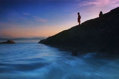 Low angle view of boy fishing white standing on rock by sea against sky during sunset