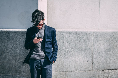 Young man using mobile phone while standing against wall