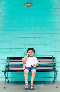 Full length portrait of boy sitting on bench talking on phone against wall