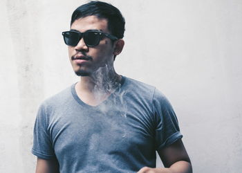 Young man smoking while standing with sunglasses by white wall