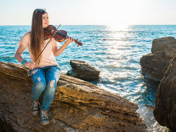 Young woman playing violin while sitting on rock in sea against sky