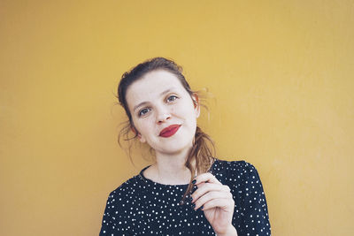 Positive young confident female model in stylish dotted dress with red lips smiling ant looking at camera while touching long brown hair against yellow background