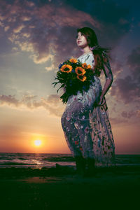 Woman standing by sea against sky during sunset