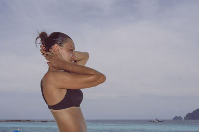 Side view of woman wearing bikini standing by sea against sky