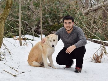 Portrait of young man smiling while crouching with dog on snow covered field