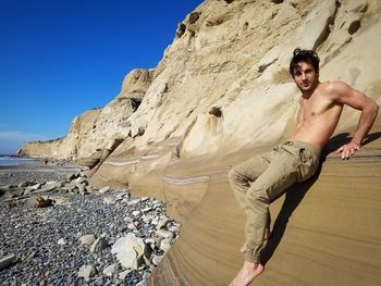 Full length of shirtless young man standing on rock formation against clear sky