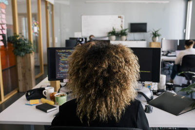 Rear view of female entrepreneur with curly hair sitting and working at desk in office