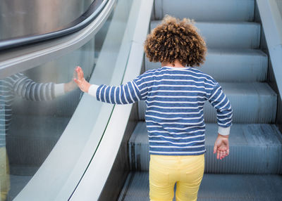 Back view of unrecognizable cute ethnic curly haired girl in trendy outfit standing on stair of escalator in city