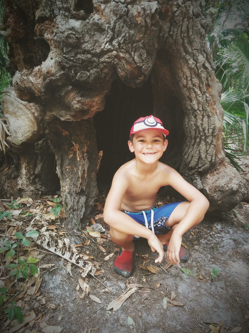 tree trunk, looking at camera, tree, one person, smiling, happiness, real people, portrait, childhood, leisure activity, front view, shirtless, outdoors, day, full length, lifestyles, nature, growth, boys