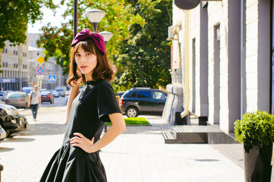 Beautiful young woman in black dress standing on sidewalk in city