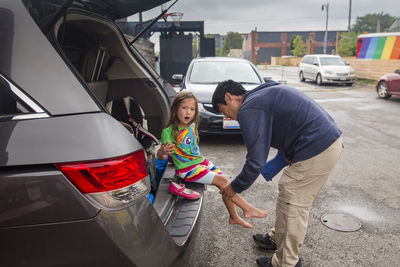 Father helping daughter in wearing shoes on car trunk