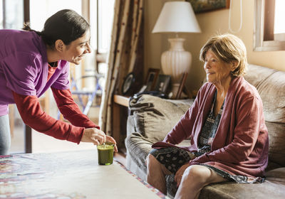 Happy caregiver stirring coffee and talking to senior woman sitting on sofa at home