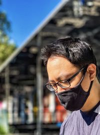 Side view of man in eyeglasses and face mask looking away against architecture, greenery and sky