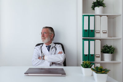 Dreamy elderly male doctor in eyeglasses with stethoscope looking away at table with tablet in hospital