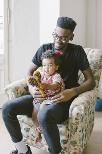 Playful baby girl playing holding maracas while sitting with father on armchair at home