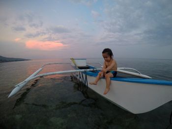 Full length of cute shirtless boy sitting on boat in sea against sky during sunset