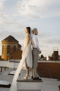 Side view of young bridge and groom standing back to back on rooftop
