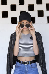 Portrait of young woman wearing sunglasses and leather jacket against wall