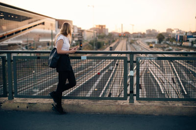 Side view of young woman standing on bridge over railroad tracks in city during sunset