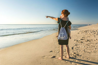 Rear view of mother pointing towards sea while carrying daughter at beach against sky