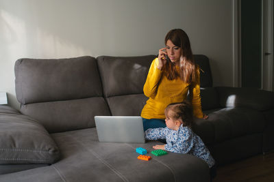 Young female freelancer having phone conversation and discussing details of project with client while working with laptop on sofa in living room with little daughter