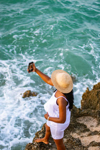 Rear view of woman standing on rock by sea holding a mobile phone